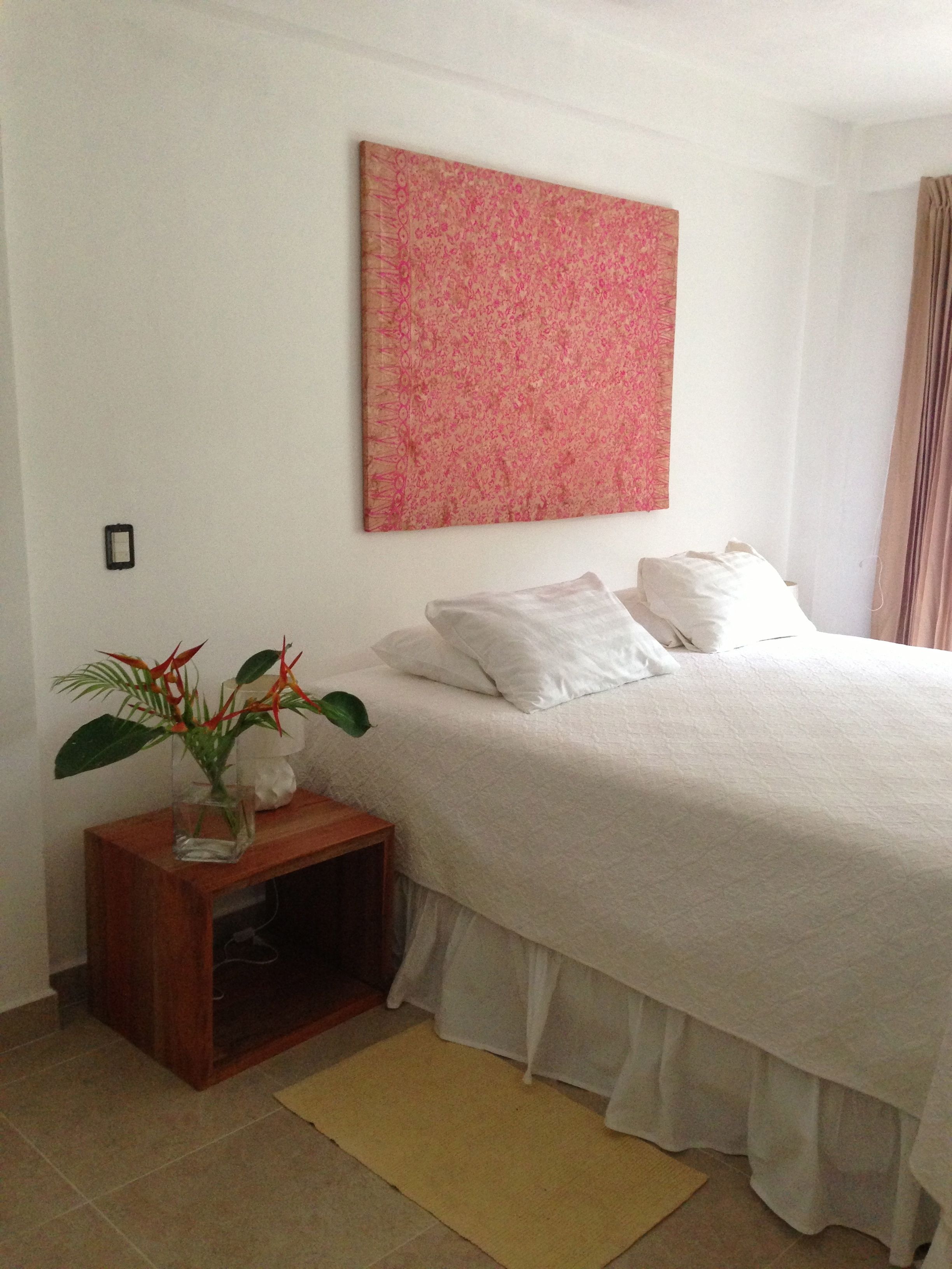 Un dormitorio con cama, una mesita de noche con una planta, una obra de arte en la pared y una ventana con cortinas.
