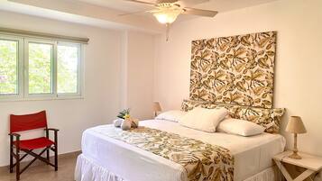 Um quarto com cama, ventilador de teto, janela, cadeira e mesa de cabeceira com luminária.