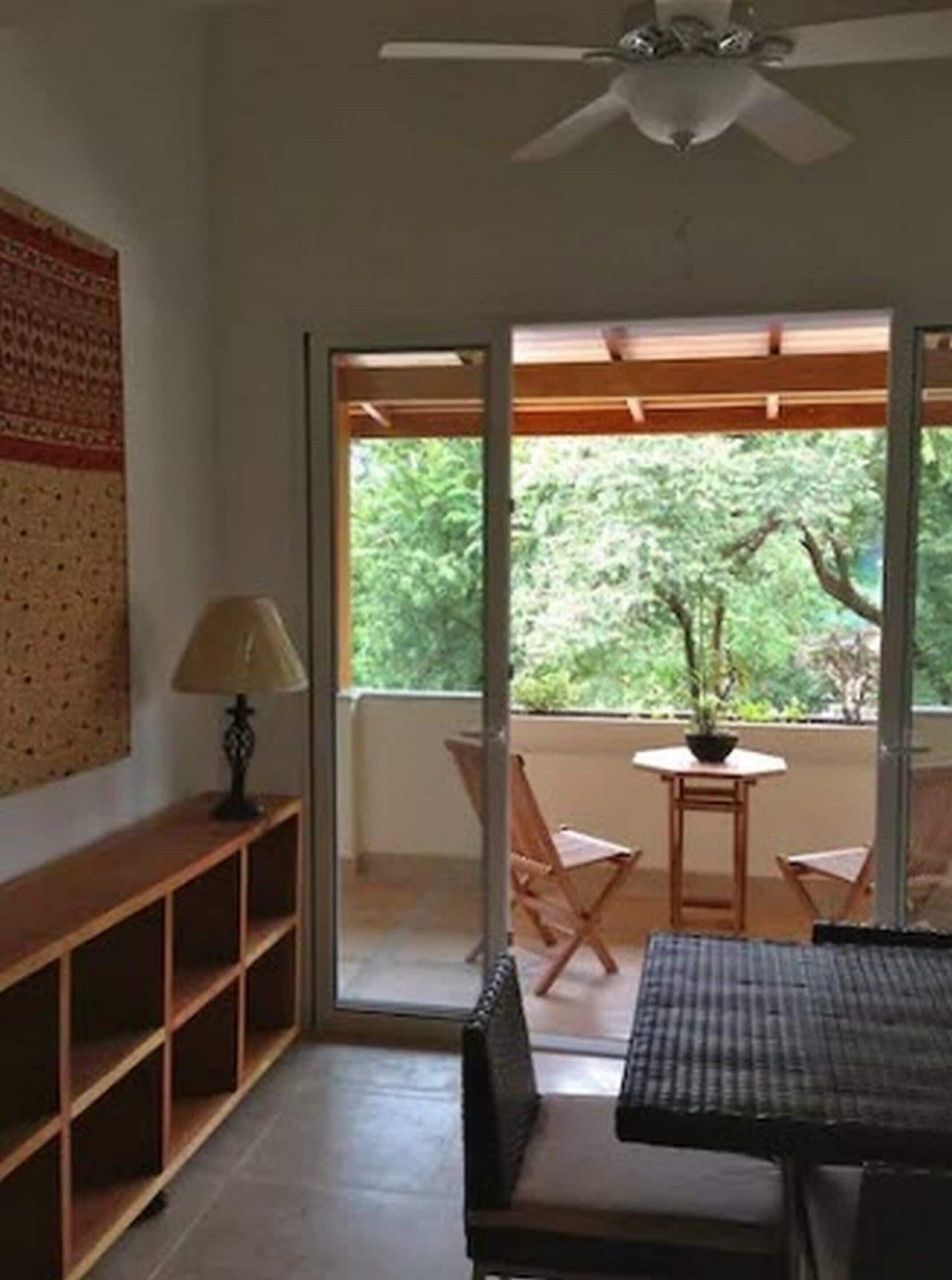 A room with a wooden bookshelf, a table with chairs, a ceiling fan, and a view of greenery outside.