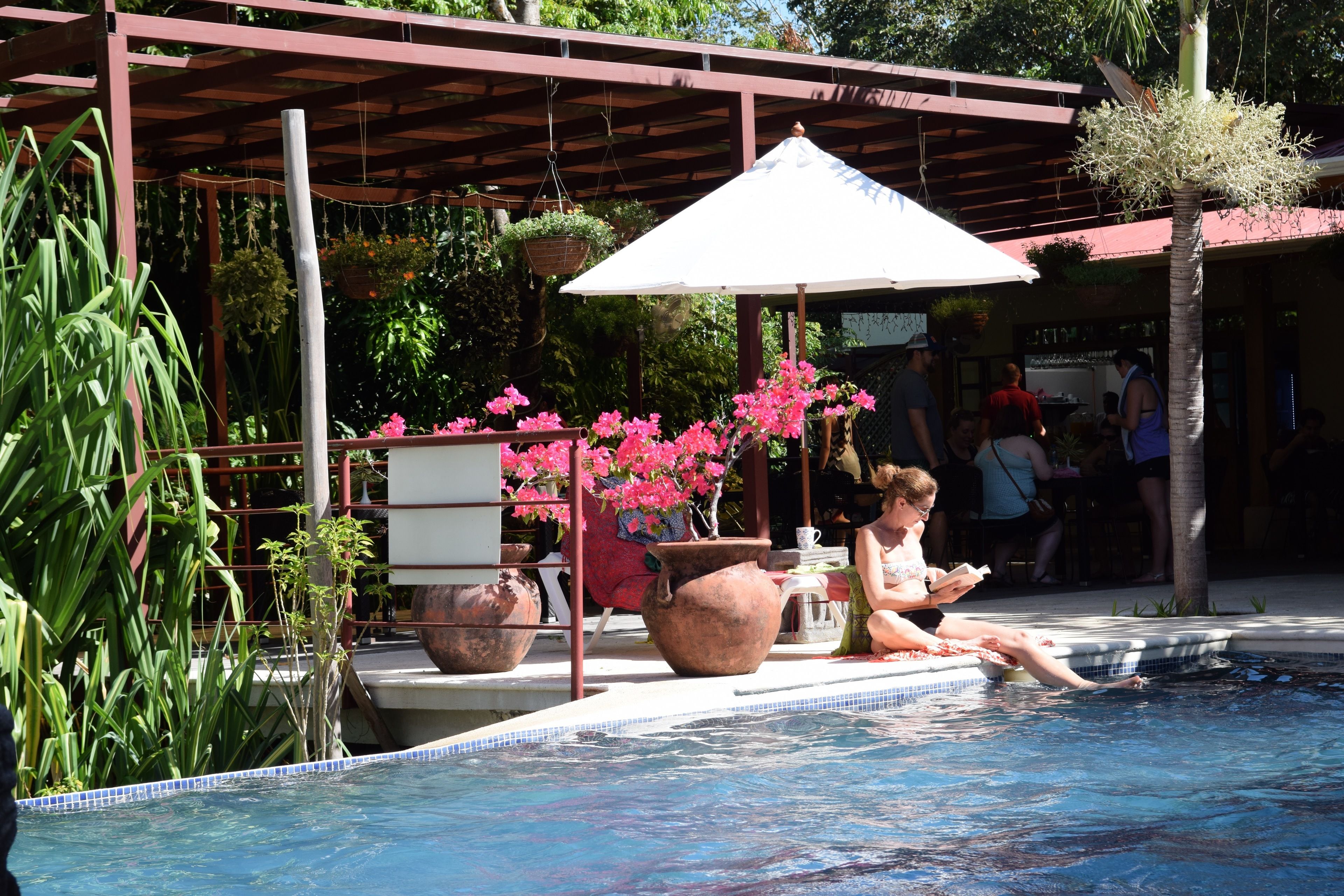 Un área de piscina con una pérgola, plantas en macetas y una persona relajándose junto a la piscina.
