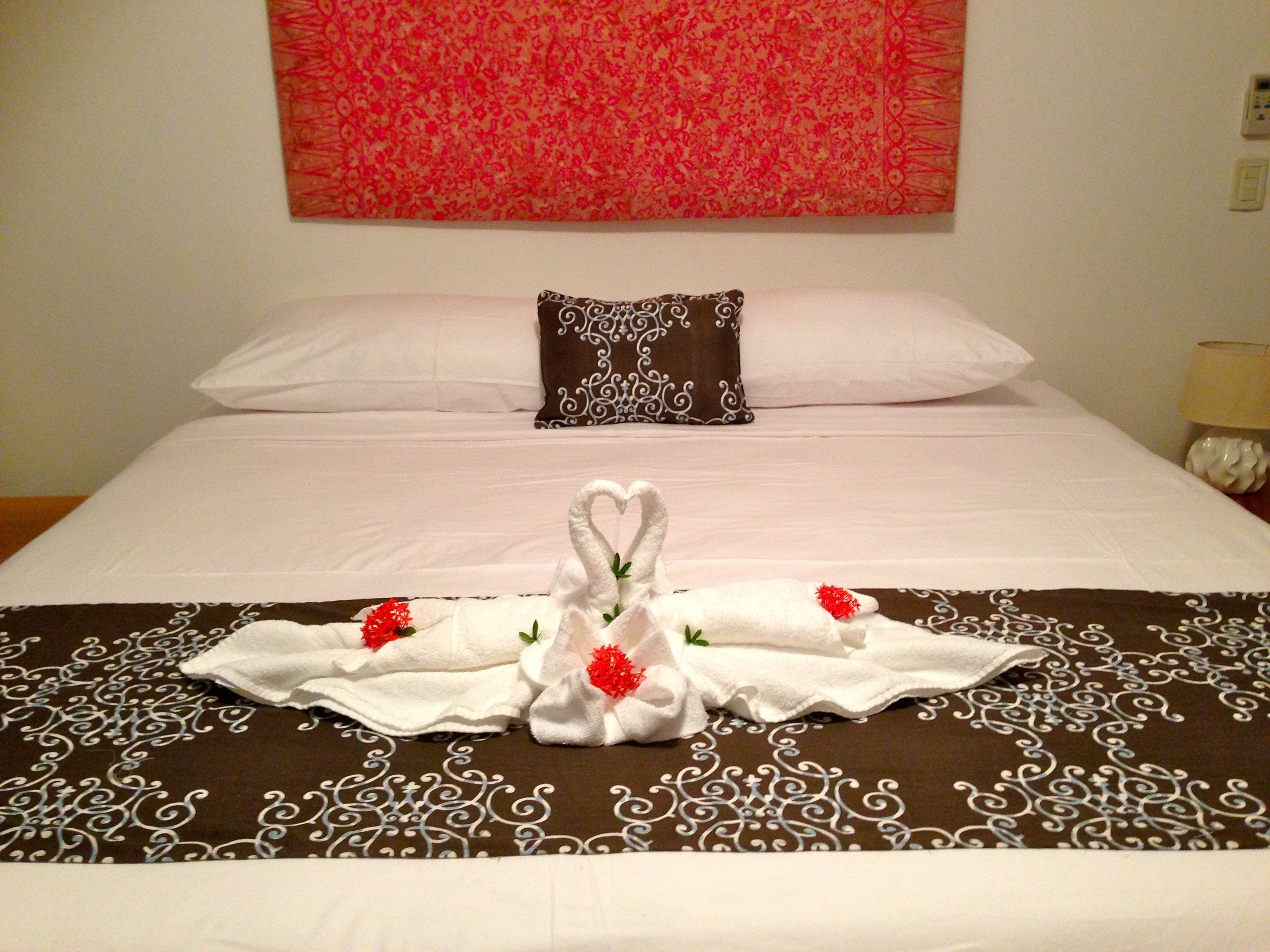 A neatly made bed with a decorative towel arrangement featuring swans and red flowers, a patterned brown and white bedspread, and a red floral tapestry on the wall.