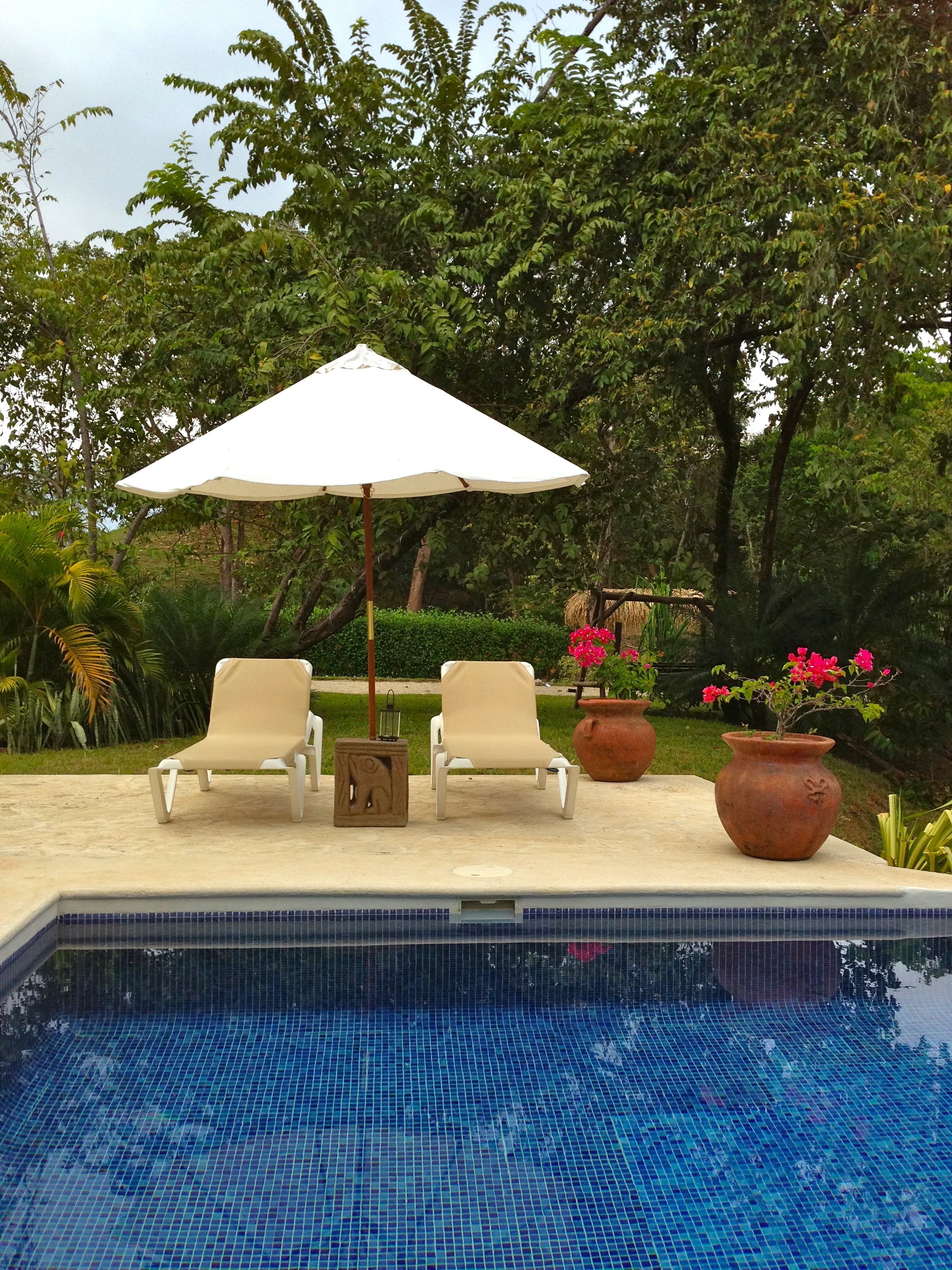 A pool area with two lounge chairs, a white umbrella, and potted plants.