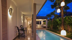 Una piscina con zona de patio, una mesa de comedor con sillas e iluminación decorativa en la pared.