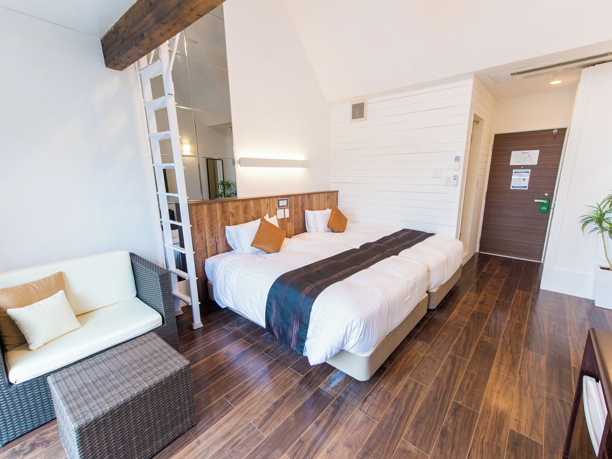 A hotel room with a large bed, a sofa, a wooden ladder, and a door with a green handle.