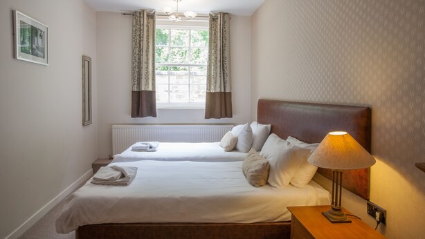 Habitación de hotel con dos camas, una mesita de noche, una lámpara, una ventana con cortinas y un radiador.