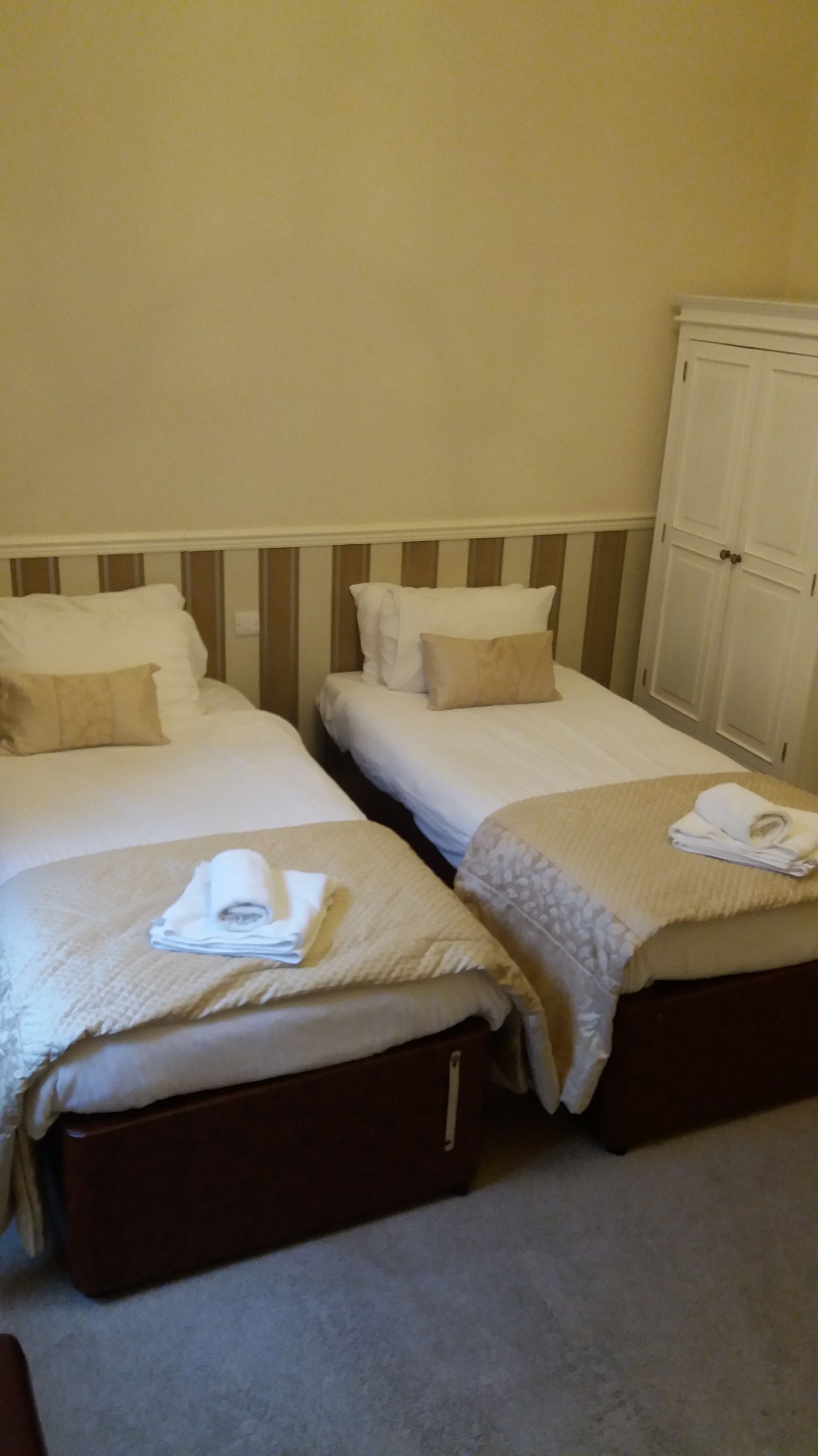 Habitación de hotel con dos camas, cada una con una almohada y una toalla doblada al lado. Se ve un armario con puerta y un interruptor de luz en la pared.