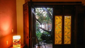 Una habitación con cama, una mesita de noche con lámpara, una silla y vistas a un jardín por una puerta abierta.