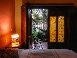 Una habitación con cama, una mesita de noche con lámpara, una silla y vistas a un jardín por una puerta abierta.