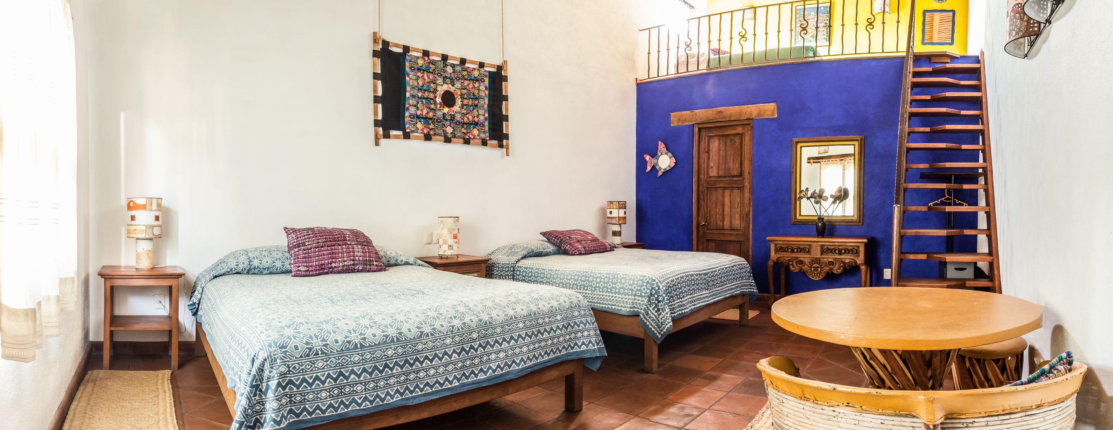 Una habitación con dos camas, una mesa de madera, un balcón y una pared azul.