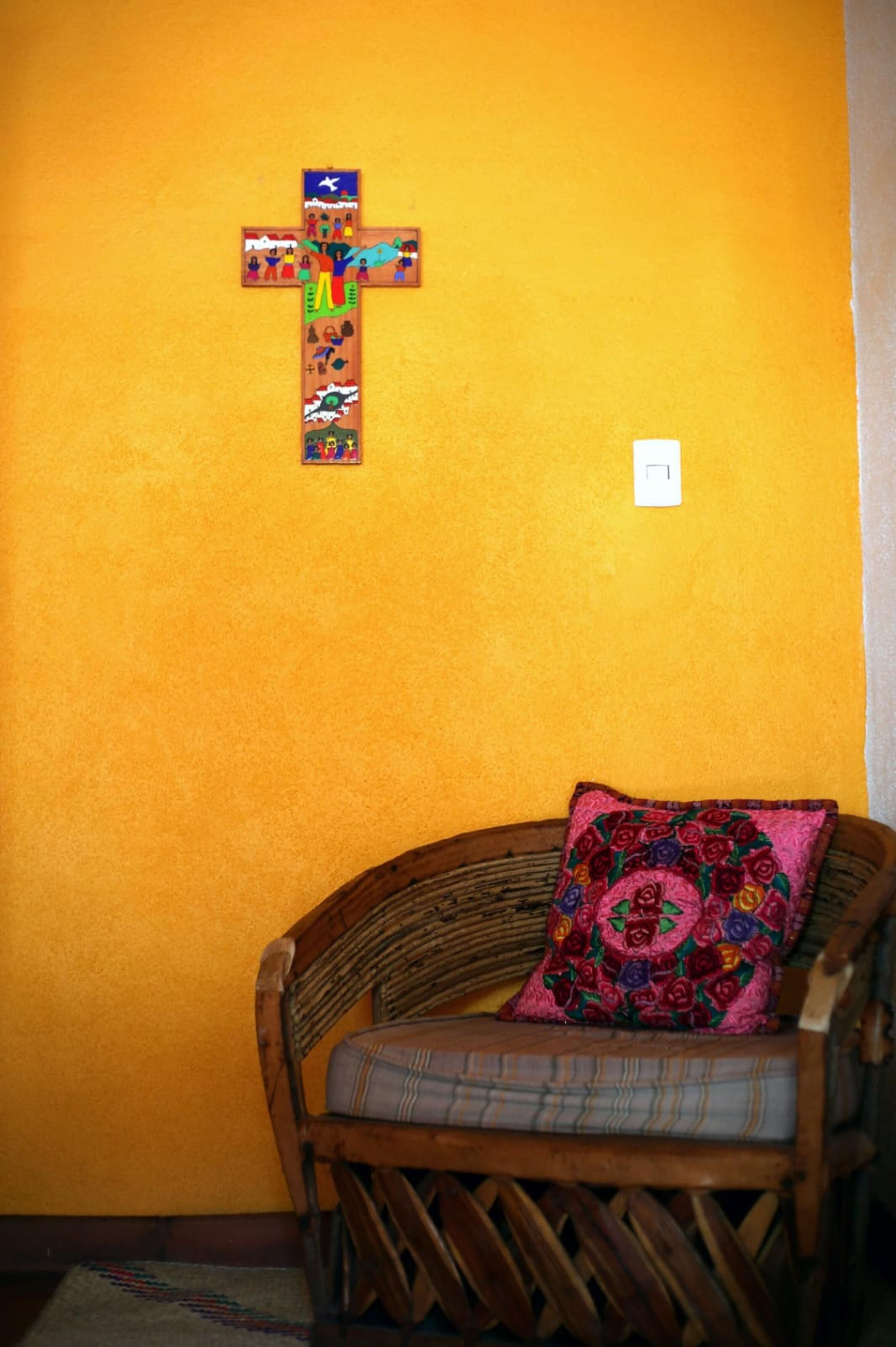 A room with a wooden chair and a colorful cross on the wall.