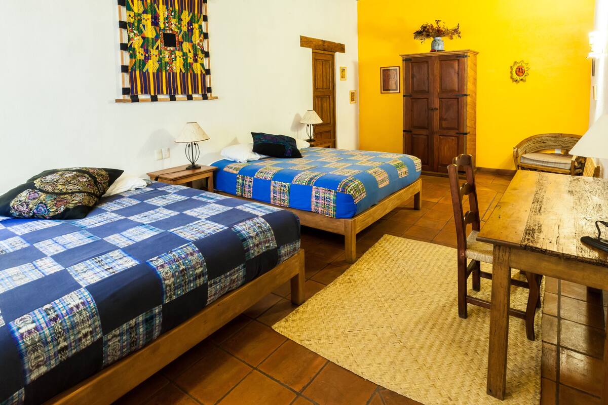 A room with two beds, a wooden table, a chair, a lamp, a cabinet, and a wall with a colorful tapestry.