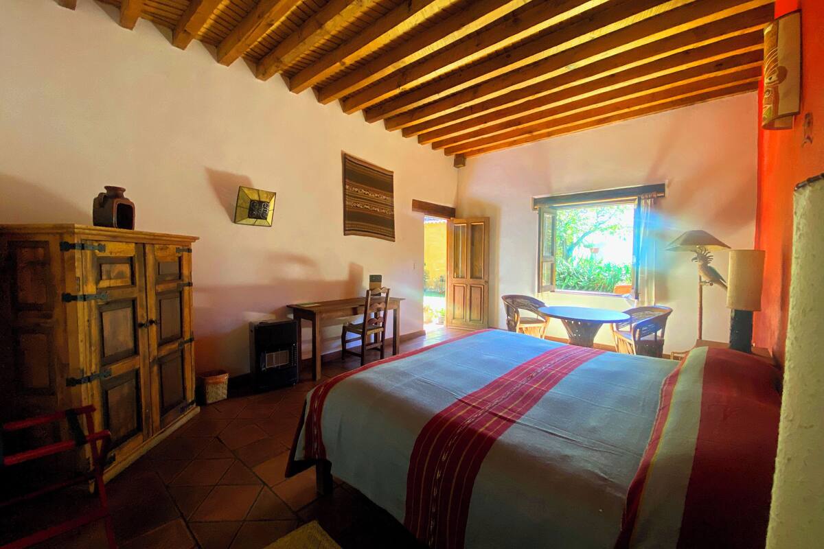 A bedroom with a wooden ceiling, a bed with a red and white bedspread, a wooden cabinet, a table, and chairs.
