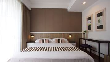 A modern hotel room with a large bed, wooden headboard, two bedside tables with lamps, a small desk, and a potted plant.