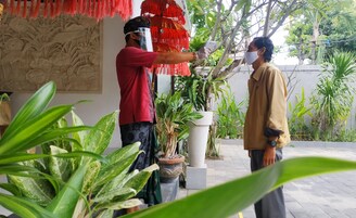 Un hombre con una camisa roja y una mujer con una camisa color beige de pie en un patio con plantas en macetas y una pared decorativa.