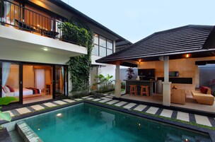 Una casa moderna con piscina, un balcón con mobiliario exterior y una cocina al aire libre cubierta.