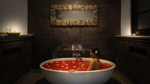 A large bathtub filled with red flowers, set in a dimly lit room with a rustic wall and a mirror above. - Agata Villas (Seminyak)