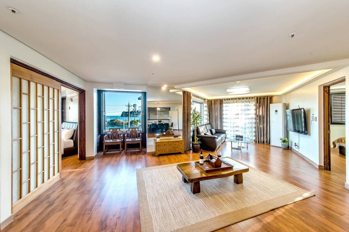 A spacious living room with wooden flooring, a large sofa, a coffee table, and a view of the cityscape through large windows.