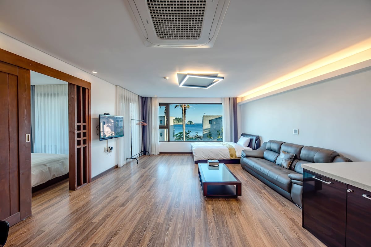 A modern hotel room with a large bed, a sofa, a glass coffee table, and a view of the sea.