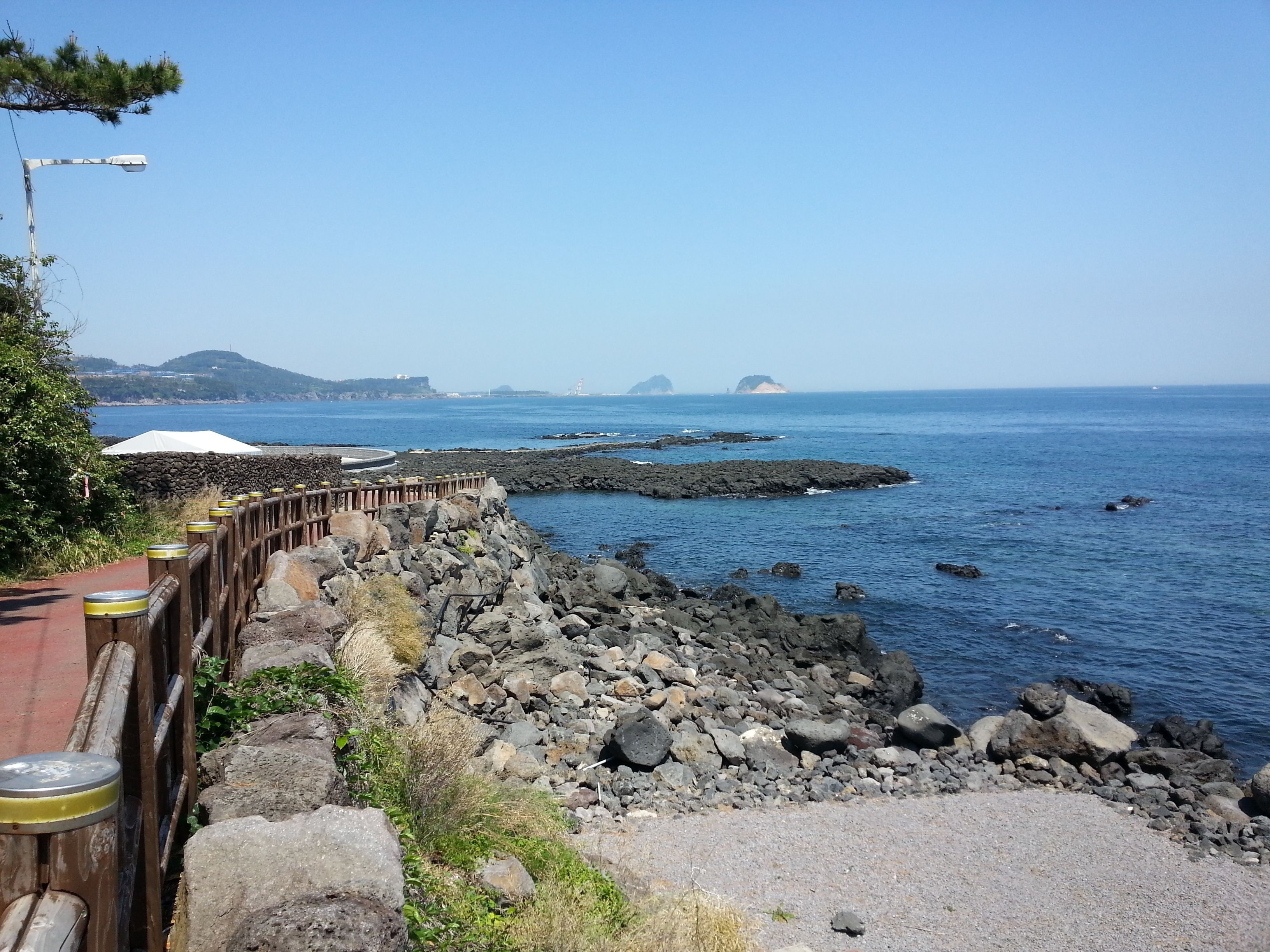 A coastal road with a wooden guardrail, rocky terrain, and a clear view of the sea and distant islands.