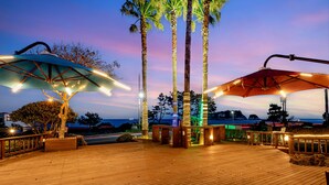 Una terraza de madera con dos sombrillas iluminadas, palmeras y vista al océano al atardecer.