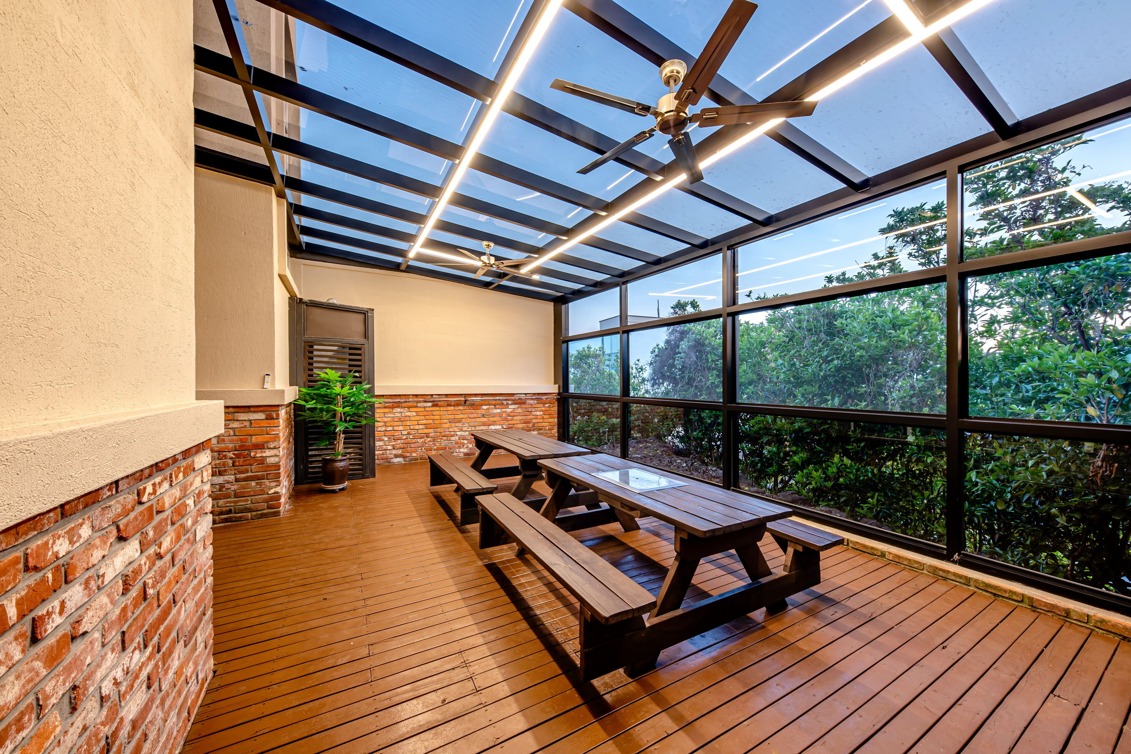 A covered patio with wooden benches and a glass ceiling, overlooking a garden.