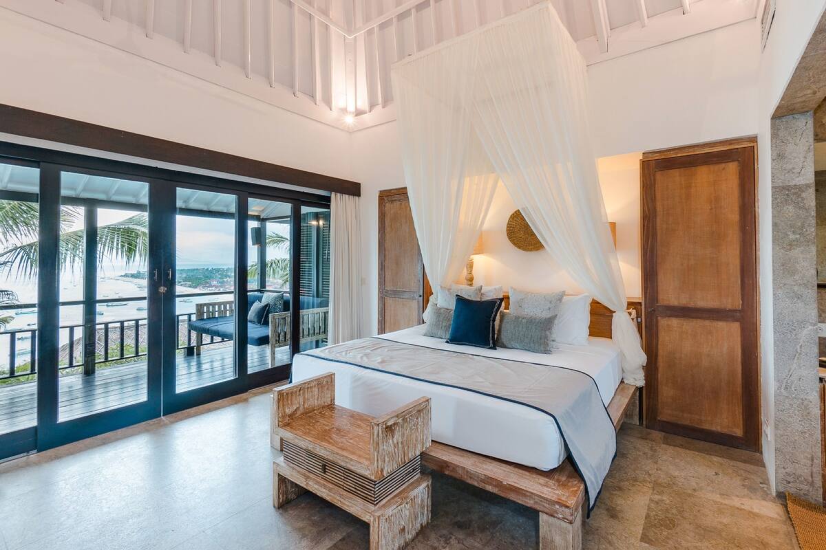 A bedroom with a large bed, a wooden bench, a nightstand, and a view of the ocean through large glass doors.