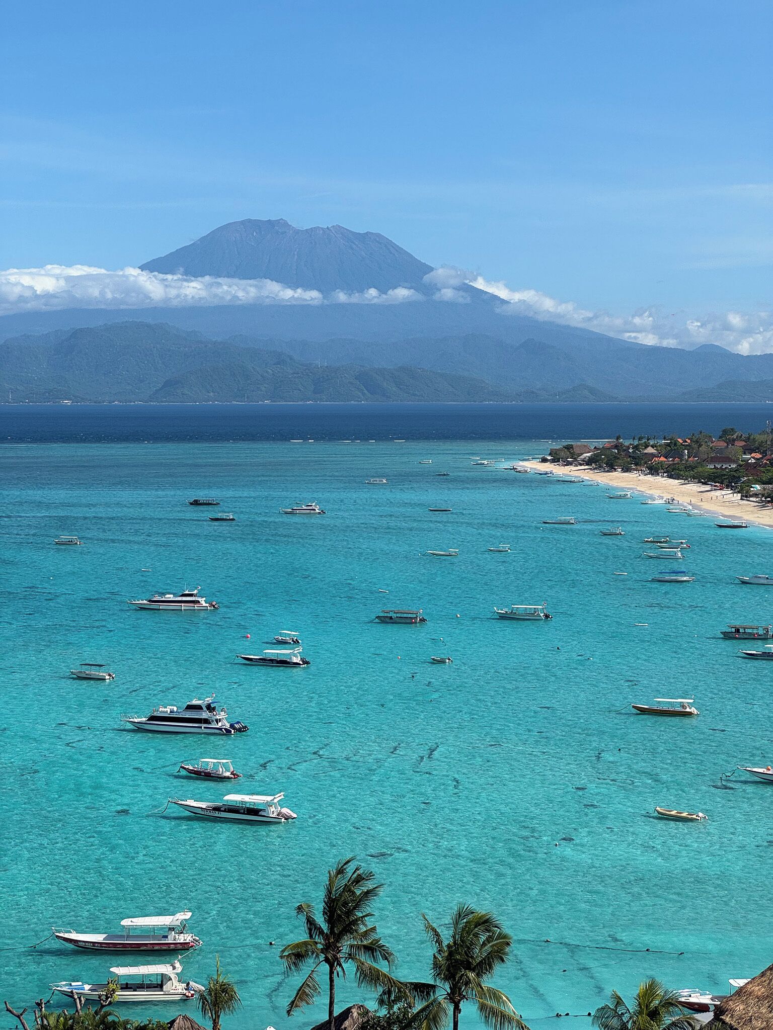 A coastal view with boats on turquoise waters, a beach, and a mountain in the background.