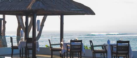 Un espace de restauration en bord de mer, doté d’un parasol en chaume, de tables, de chaises et offrant une vue sur l’océan.