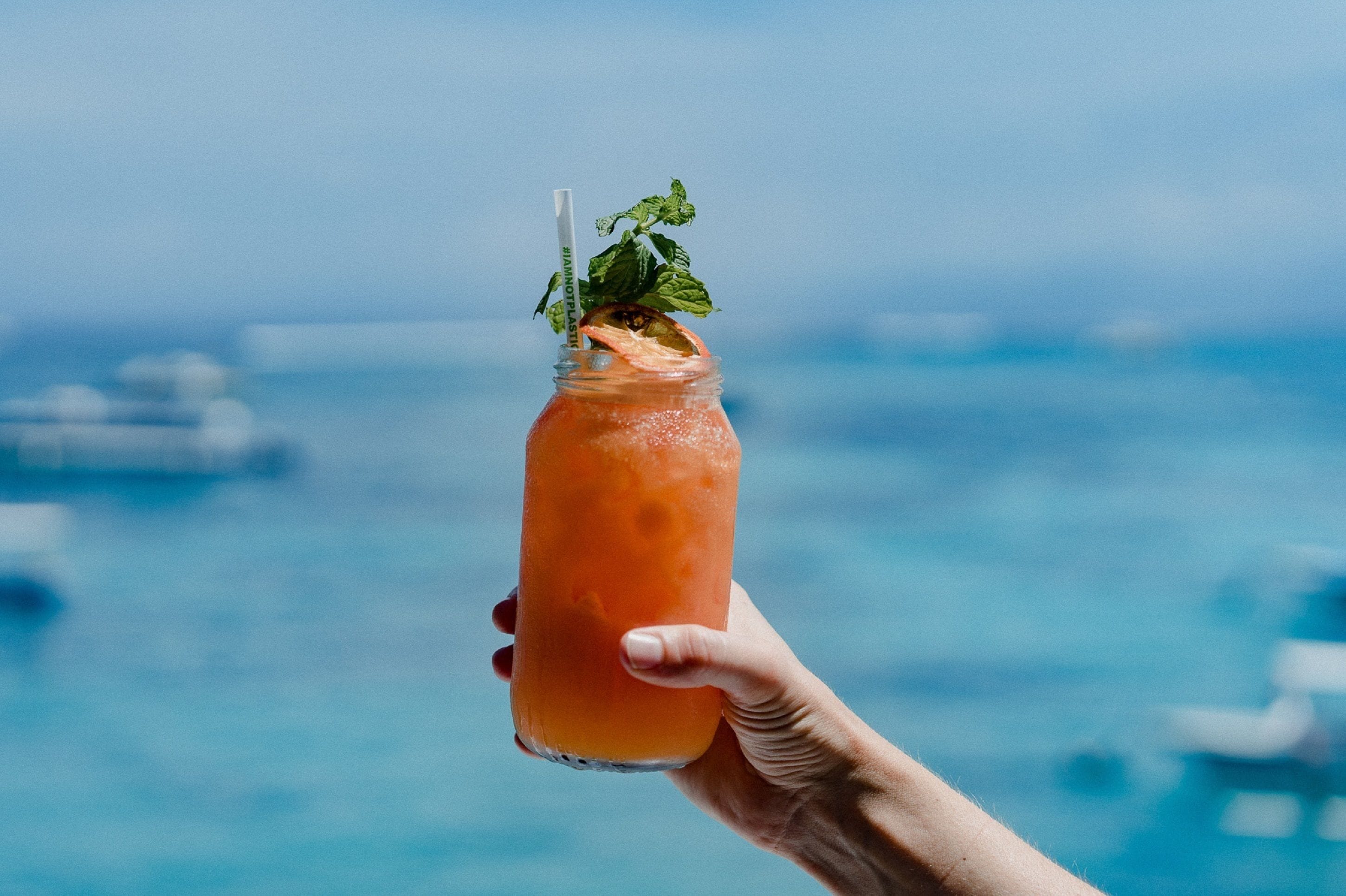 A hand holding a glass of orange beverage with a mint garnish, set against a blurred background of a body of water and boats.
