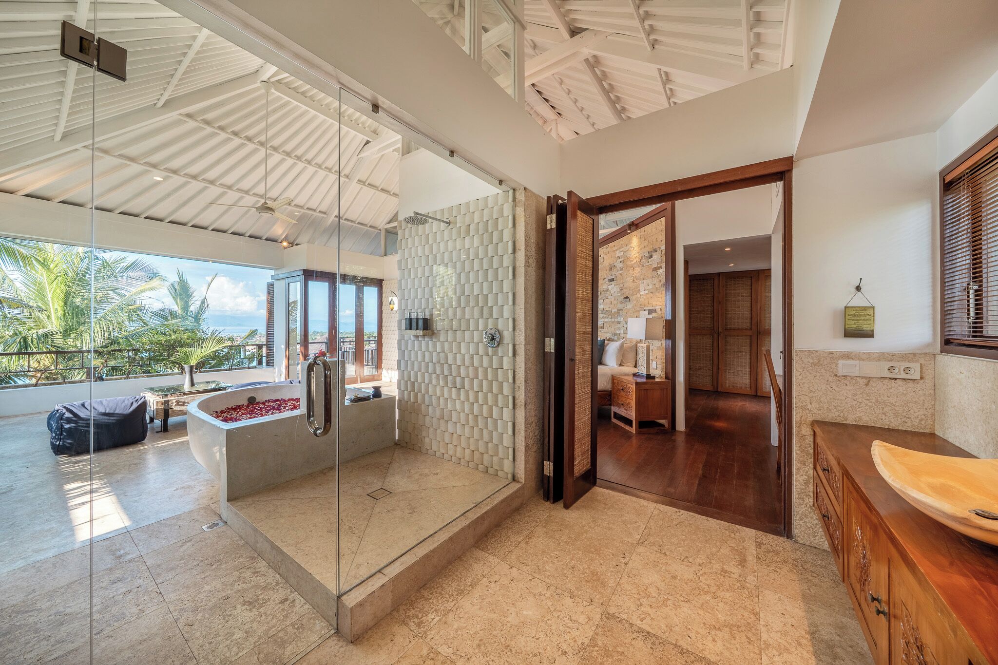 A modern bathroom with a glass-enclosed shower, a bathtub with red petals, and a view of the ocean.
