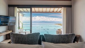 Ein Hotelzimmer mit großem Fenster und Blick auf das Meer und Boote. Es gibt einen Fernseher auf einem Standfuß, ein Bett mit Kissen und einen kleinen Tisch mit einer Pflanze.