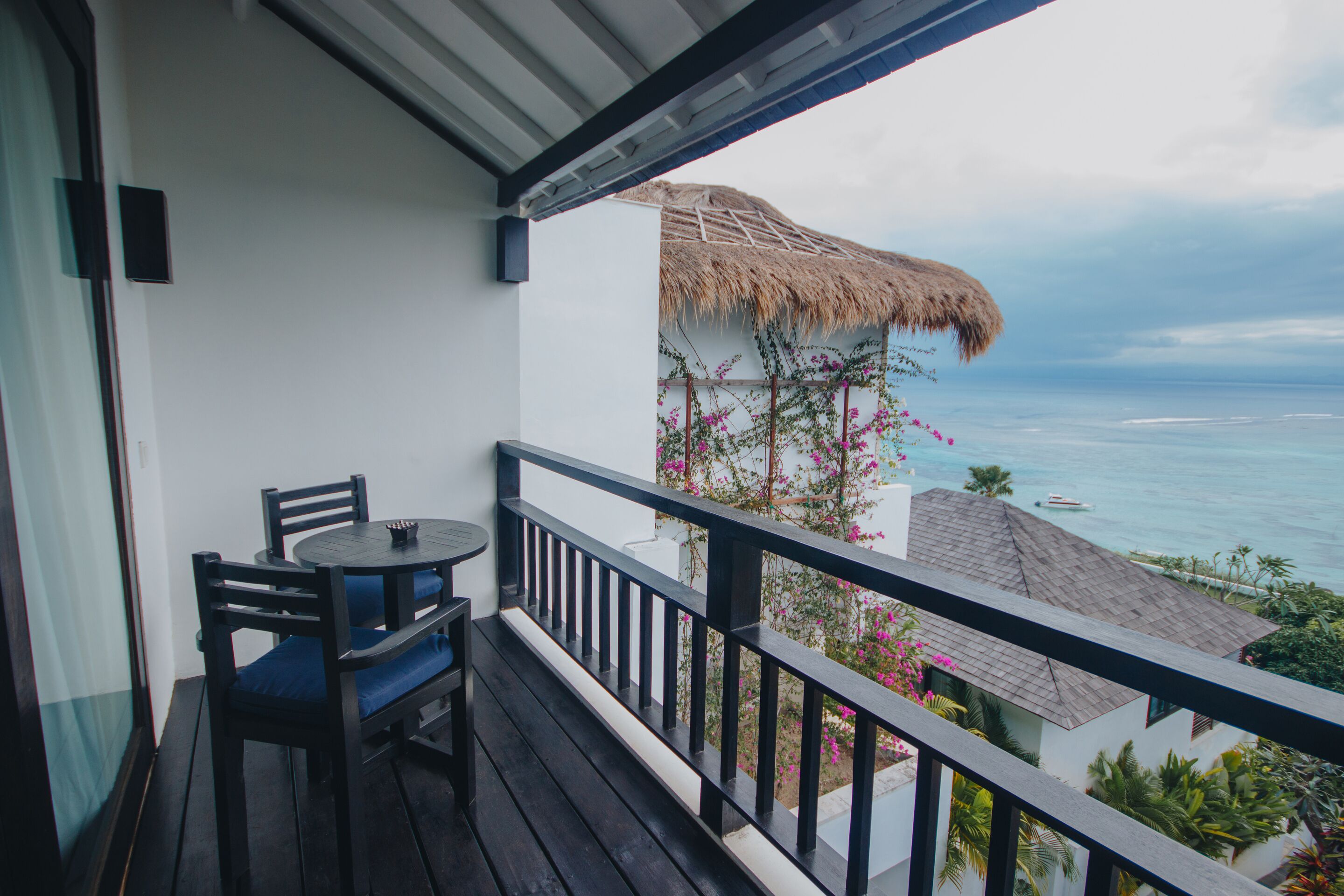 Un balcón con mesa y sillas, con vista a una playa que incluye una estructura de techo de paja y flores rosadas.