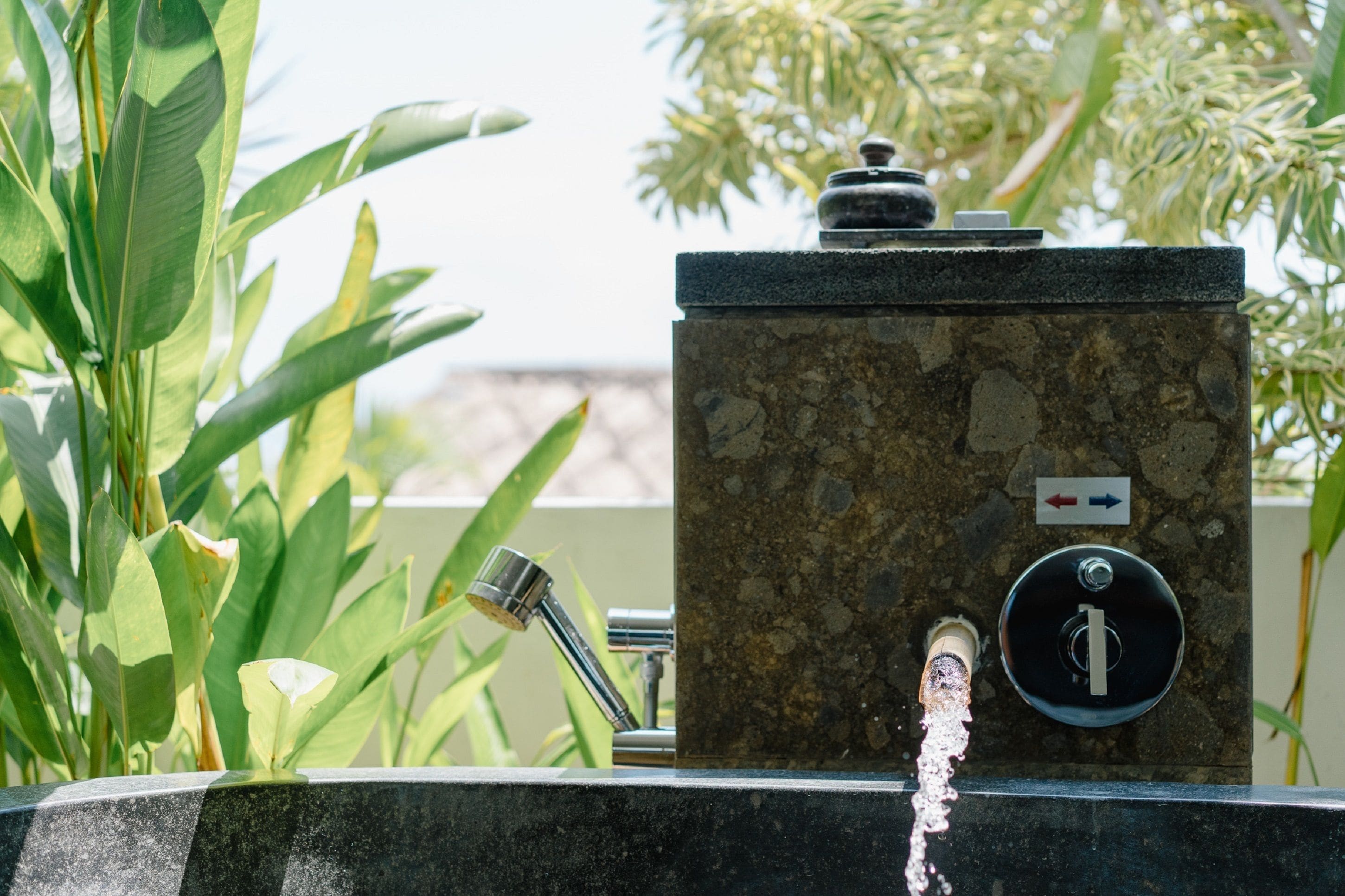 A water fountain with a stone base and a metal spout, surrounded by green foliage.