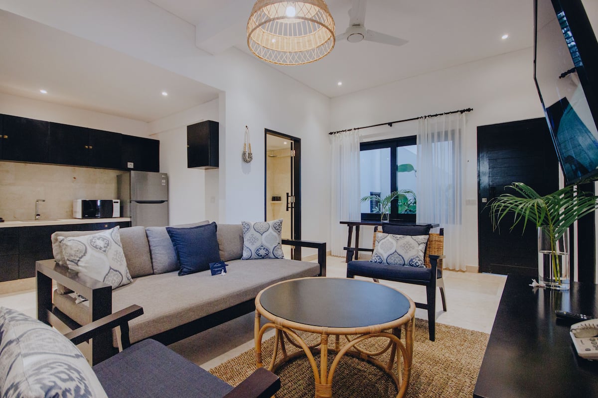A modern living room with a sofa, chairs, a round table, and a kitchen area in the background.