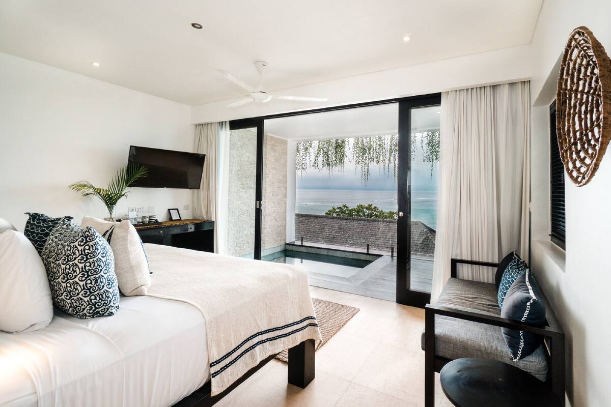 A bedroom with a bed, a TV, a desk, and a view of the sea through a glass door.