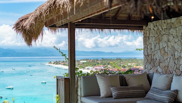 Ein Zimmer mit Steinwänden und einer gepolsterten Sitzecke mit Blick auf die Küste, Boote und das entfernte Land.