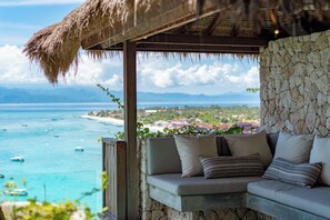 A stone-walled room with a cushioned seating area overlooking a coastal view with boats and distant land. - Batu Karang Lembongan Resort & Spa (Lembongan Island)