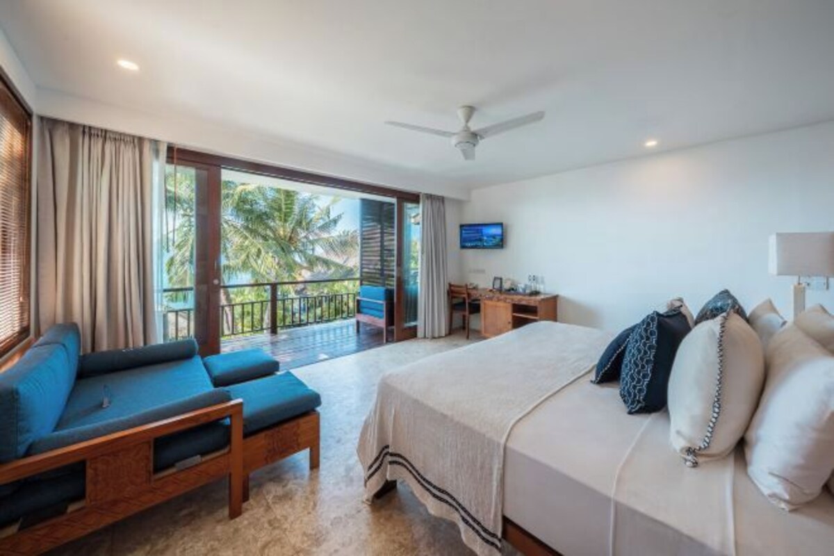 Habitación de hotel con cama, sofá, escritorio y balcón con vistas a palmeras.