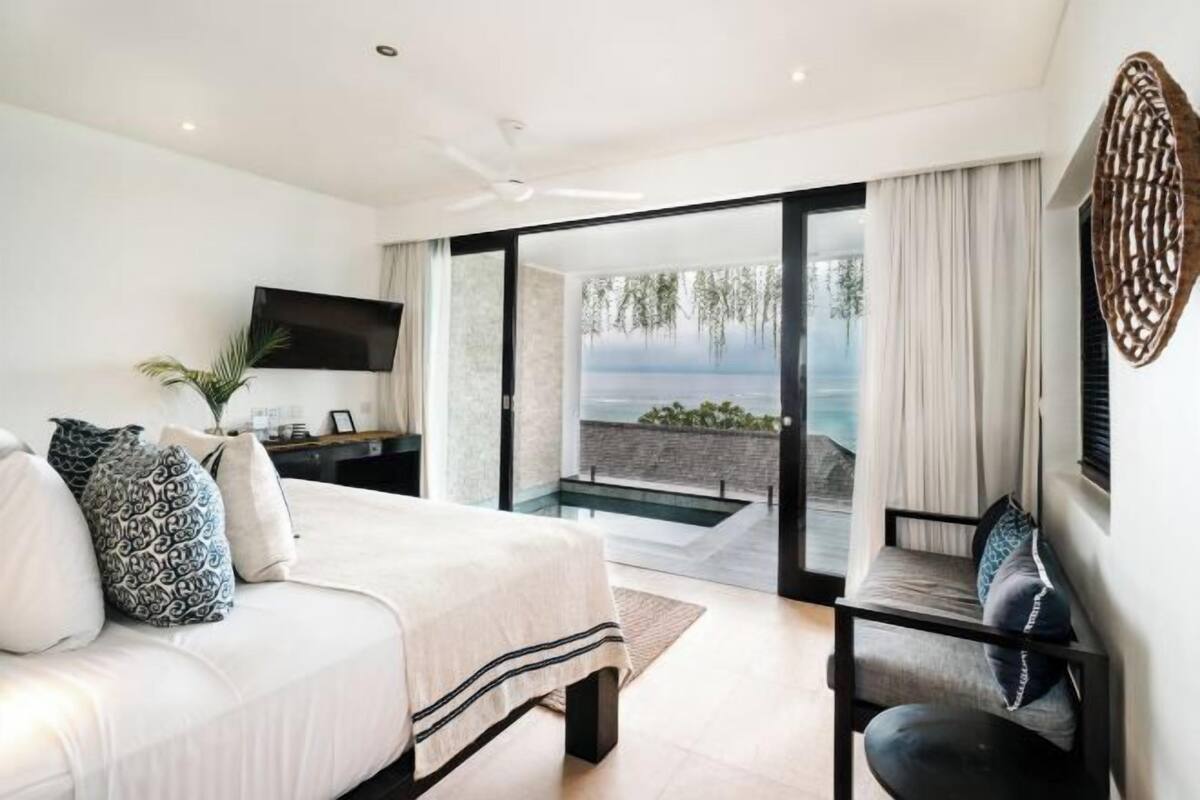 A bedroom with a bed, a TV, a sofa, and a view of the sea through a glass door.