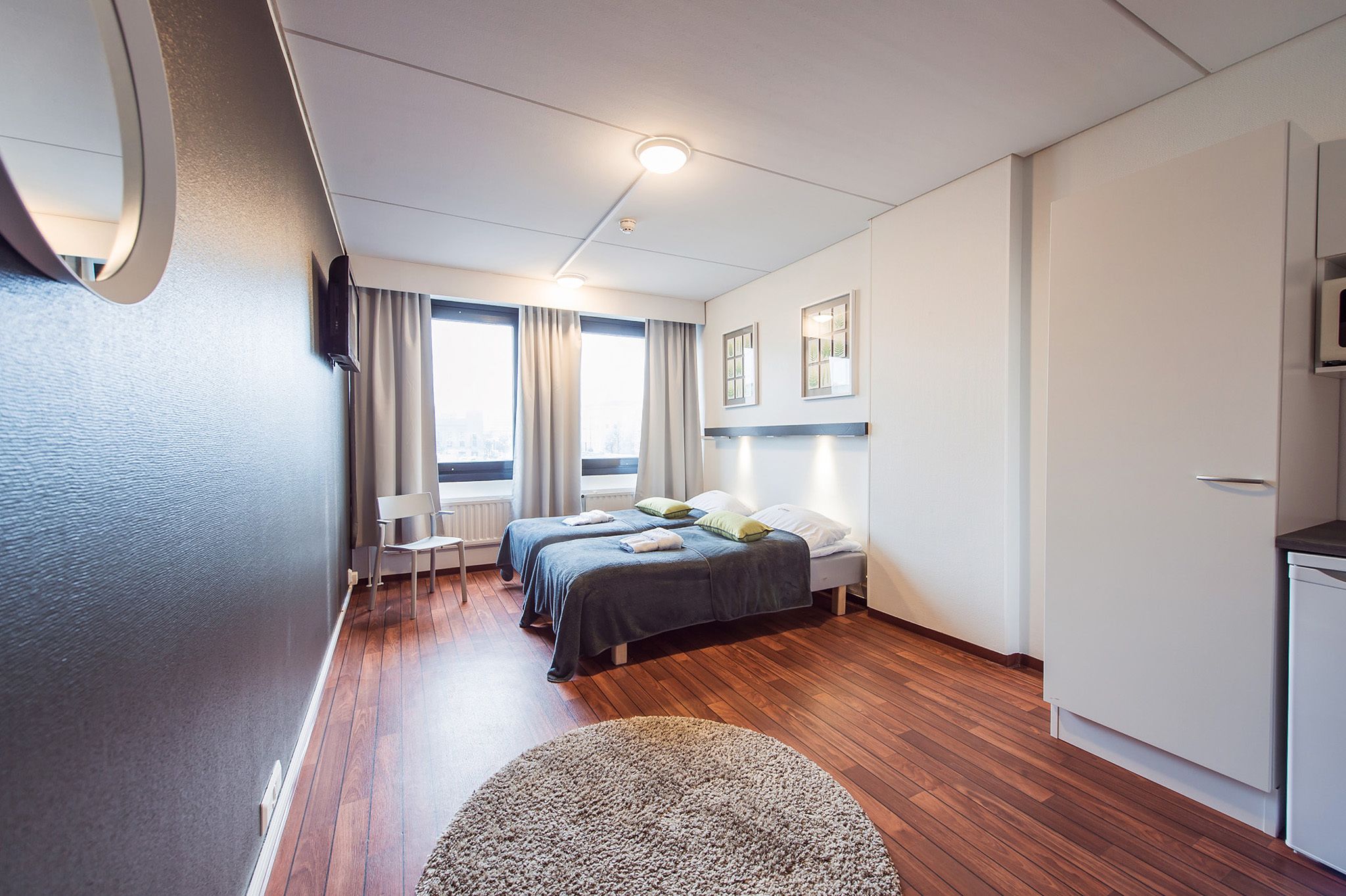 Ein modernes Hotelzimmer mit Holzboden, zwei Betten, einem Schreibtisch, einem Sessel und Blick auf die Stadt durch große Fenster.