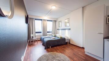 Una habitación de hotel moderna con suelo de madera, dos camas, un escritorio, una silla y vistas a la ciudad a través de amplios ventanales.
