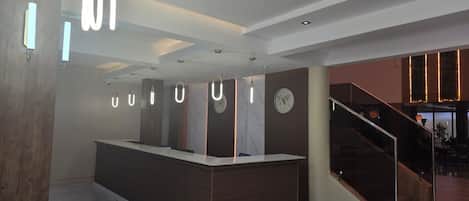 A modern reception area with a central desk, a clock, and a staircase leading to an upper level.