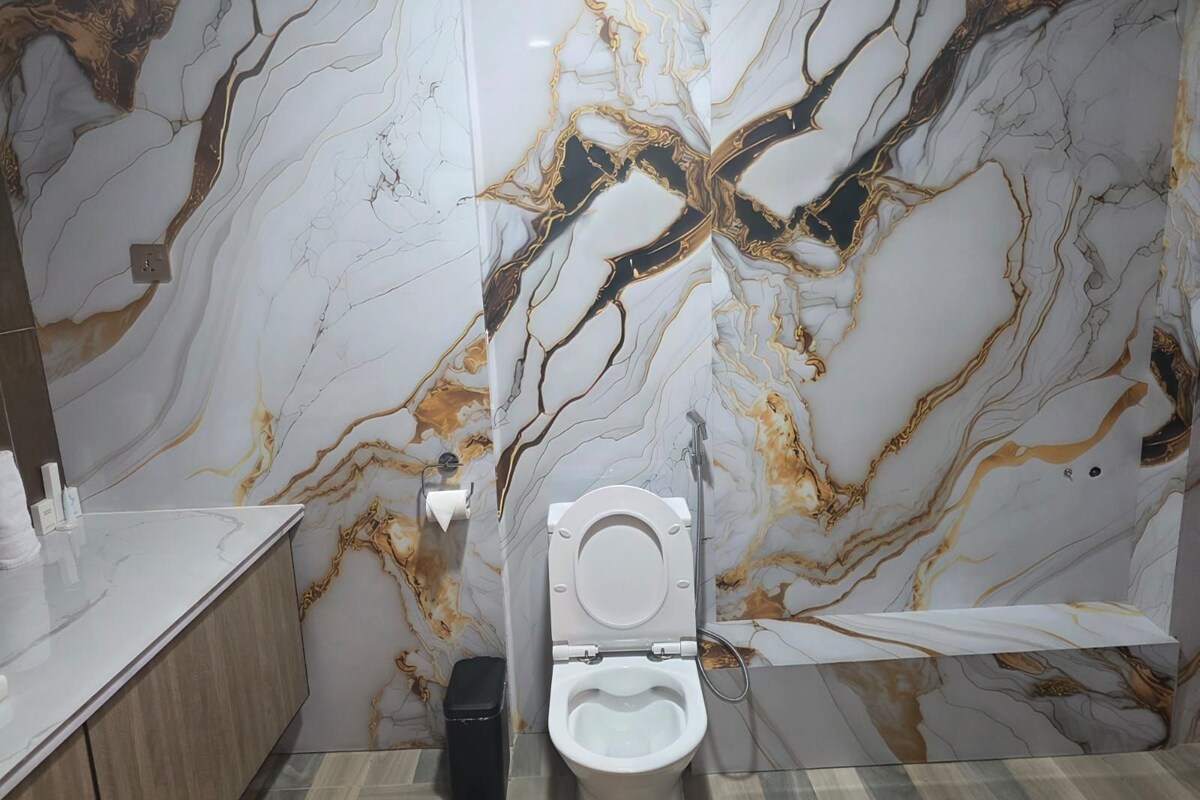 A bathroom with a marble wall, a toilet, a trash bin, and a sink with toiletries.