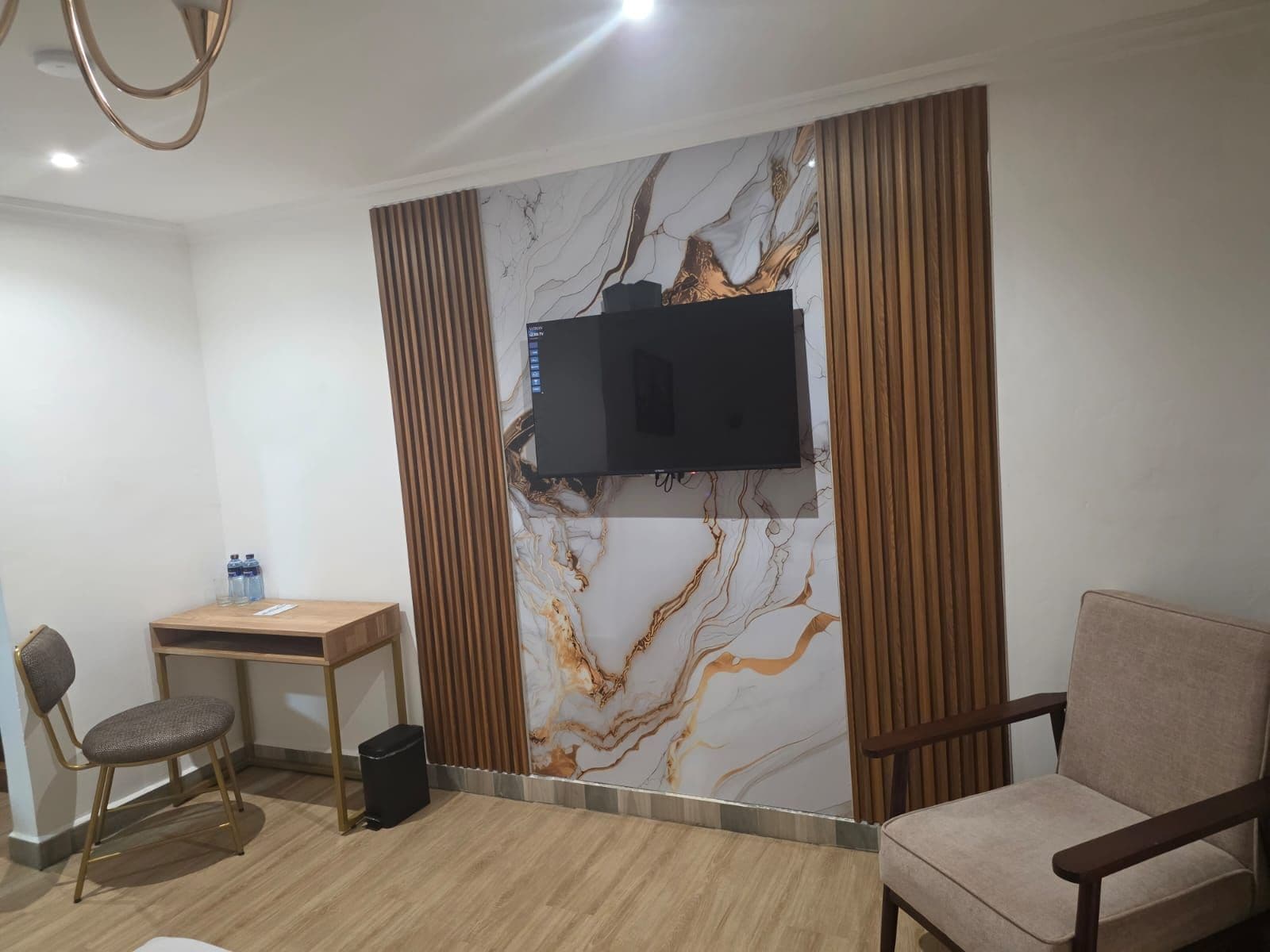 Habitación de hotel con televisor de pantalla plana montado en una pared con diseño de mármol, un escritorio de madera, dos sillas y un sillón con cojín.