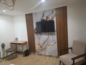 A hotel room with a flat-screen TV mounted on a marble-patterned wall, a wooden desk, two chairs, and a cushioned armchair. - Park Place Hotel (Nairobi)