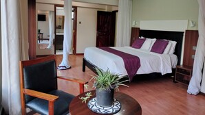 A hotel room with a large bed, a bedside table, a chair, a small table with a plant, and a view of a bathroom and hallway. - Park Place Hotel (Nairobi)