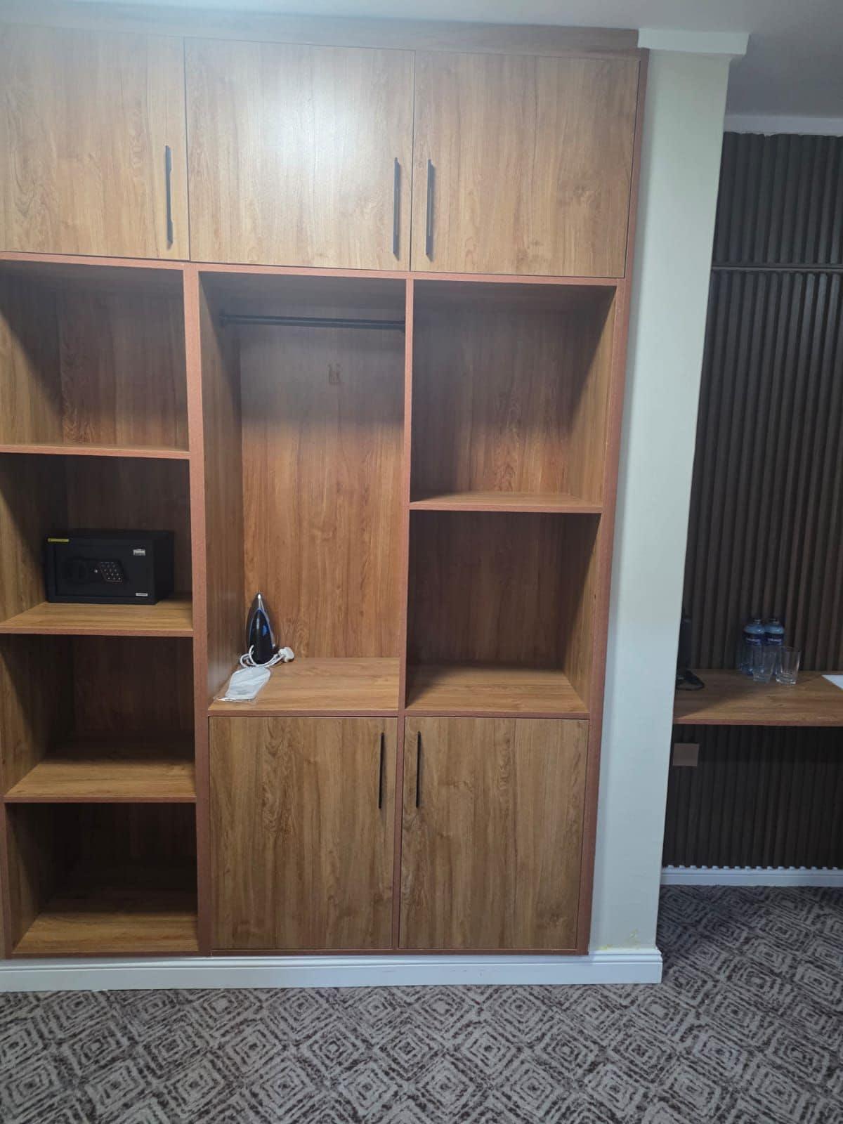 Un armario de madera con varios compartimentos, una pequeña repisa con un dispositivo negro y una mascarilla en la repisa inferior.