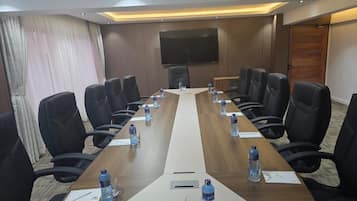 Un salón de conferencias con una larga mesa, sillas negras, botellas de agua y un televisor de pantalla plana en la pared.
