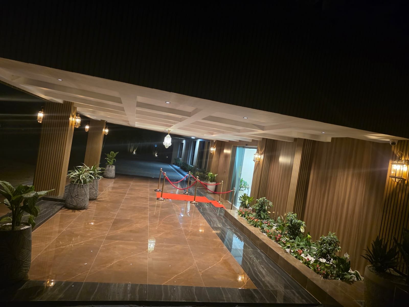 A hotel lobby with a marble floor, potted plants, and a red carpet leading to the entrance.