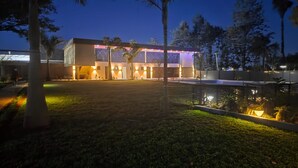 Um edifício moderno com telhado plano e janelas amplas, cercado por um gramado bem iluminado e palmeiras.